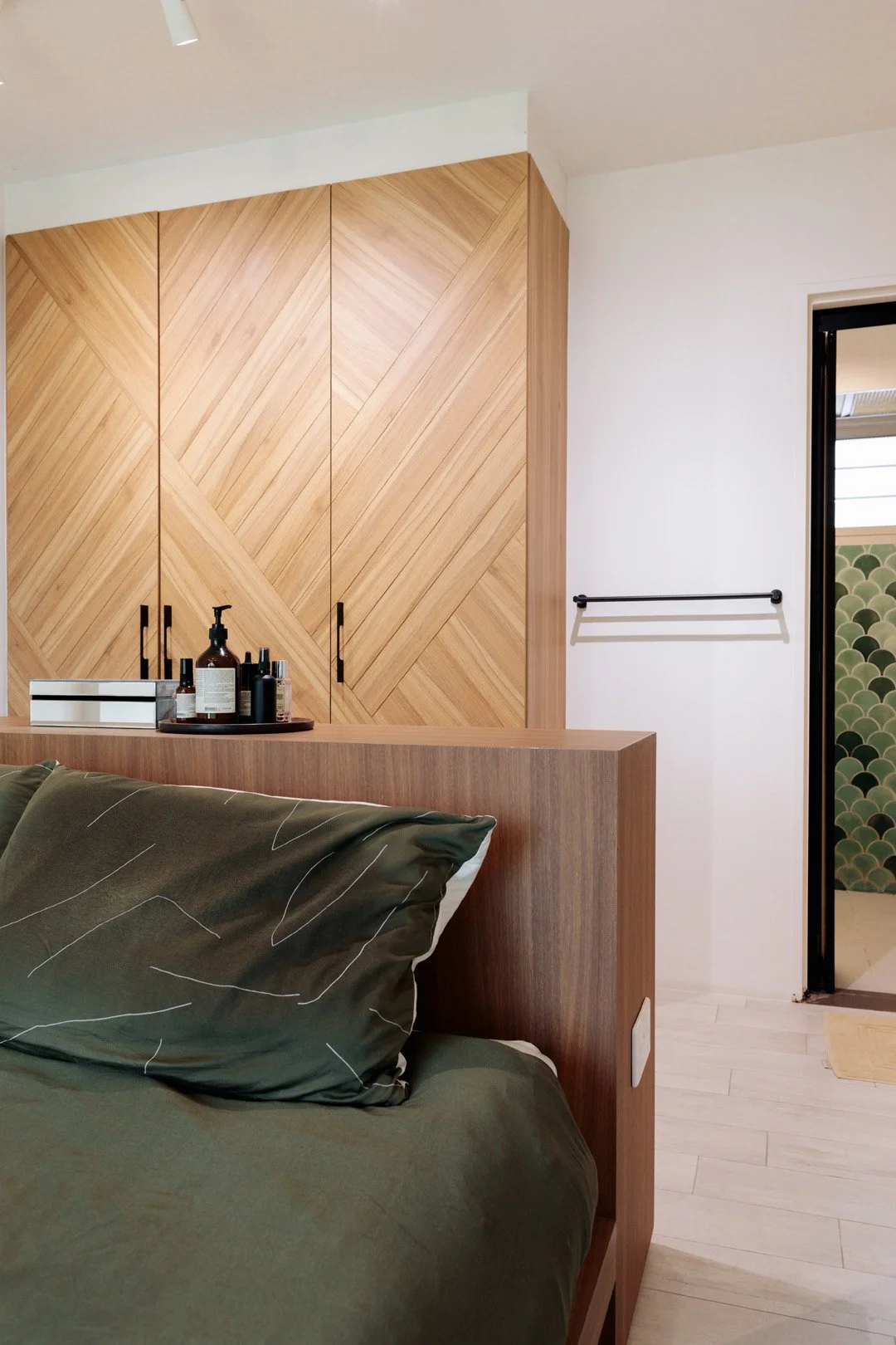 A bedroom with a wooden headboard and wardrobe. The bed has a dark green pillow and matching bedsheet. A tray with various bottles and a box are placed on a surface behind the bed. To the right, there's an open doorway showing a patterned green and b