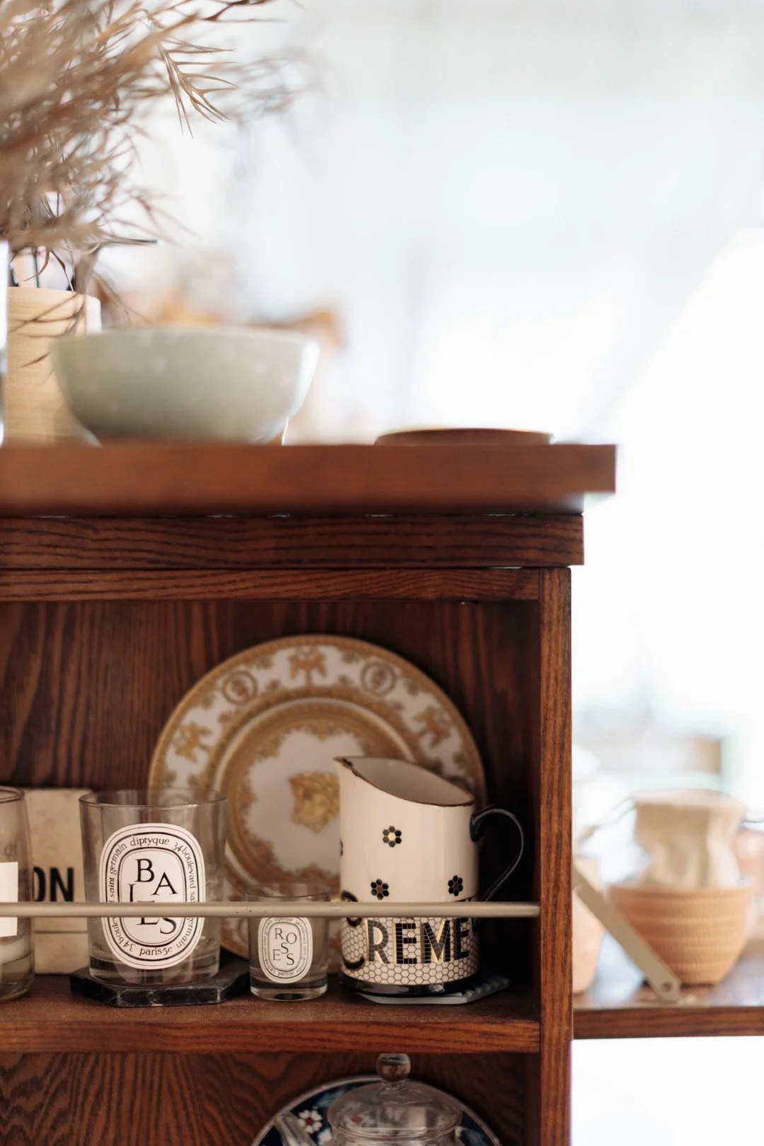 Close-up of a wooden shelf with ceramic and glassware, including a cup with 'CREME' written on it, decorative plates, and glasses, with additional items blurred in the background.