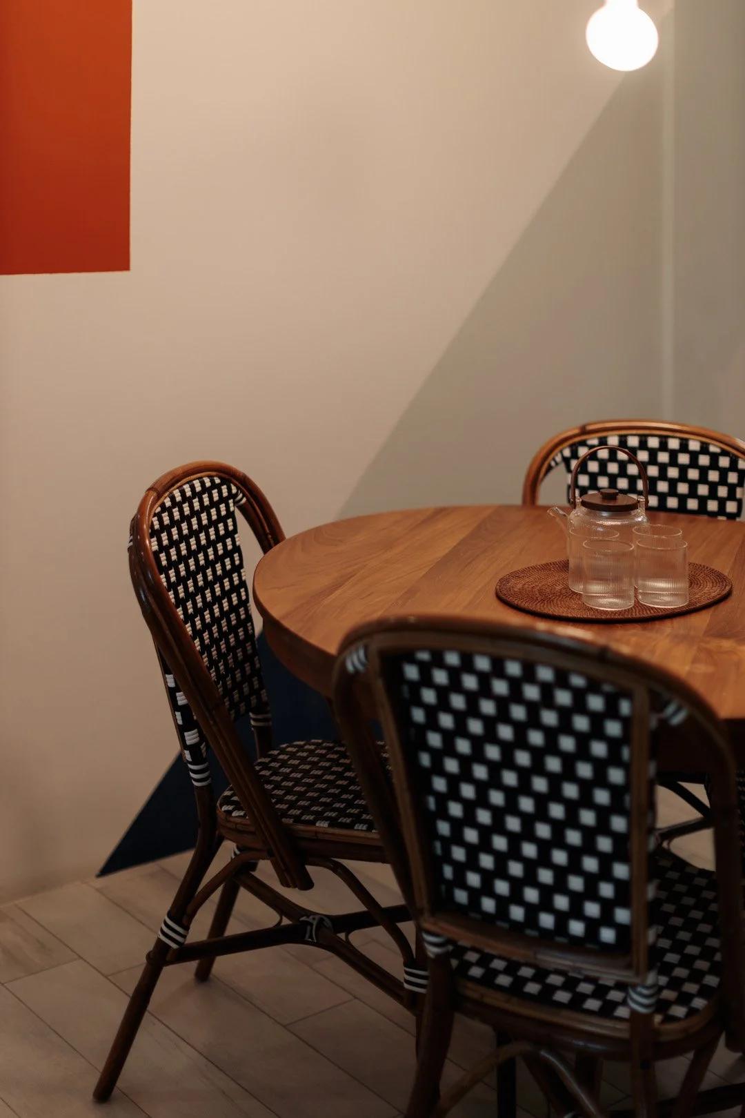 A wooden dining table with a glass pitcher and three glasses on a woven placemat. Four rattan chairs with black and white checkered cushions surround the table. A white wall with a red square and a hanging light fixture are visible in the background.