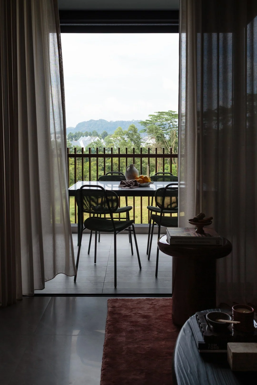 View from inside a room looking out onto a balcony with a table and four black chairs, overlooking a green landscape with trees and mountains in the distance.