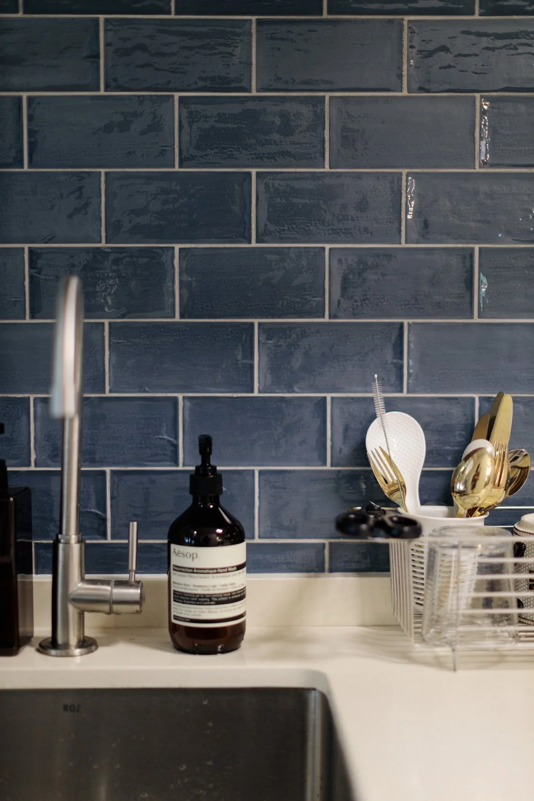Kitchen sink area with a blue subway tile backsplash, a soap dispenser labeled 'Aesop', a dish rack holding gold and black utensils, a white spatula, and a black pair of scissors.