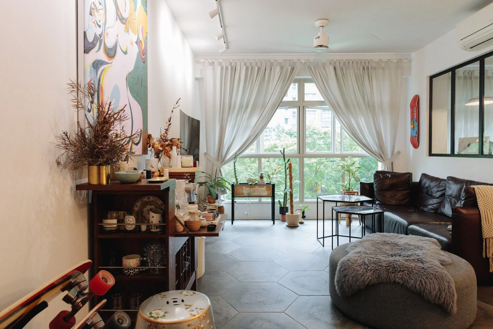 Living room with a large window and white curtains, a brown leather sofa, a round gray ottoman with a furry blanket, potted plants near the window, a wooden shelf with dishes and decorations, a wall painting, and small tables.
