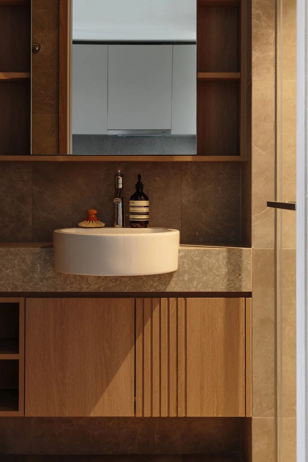 A bathroom sink with a bottle of soap or lotion and a small brush on the counter. A mirror above the sink reflects a part of a white kitchen or laundry room.
