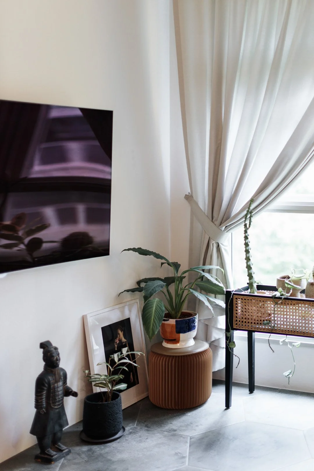 Living room corner with a wall-mounted TV, potted plants, a framed photo, and decorative statues near a large window with beige curtains.