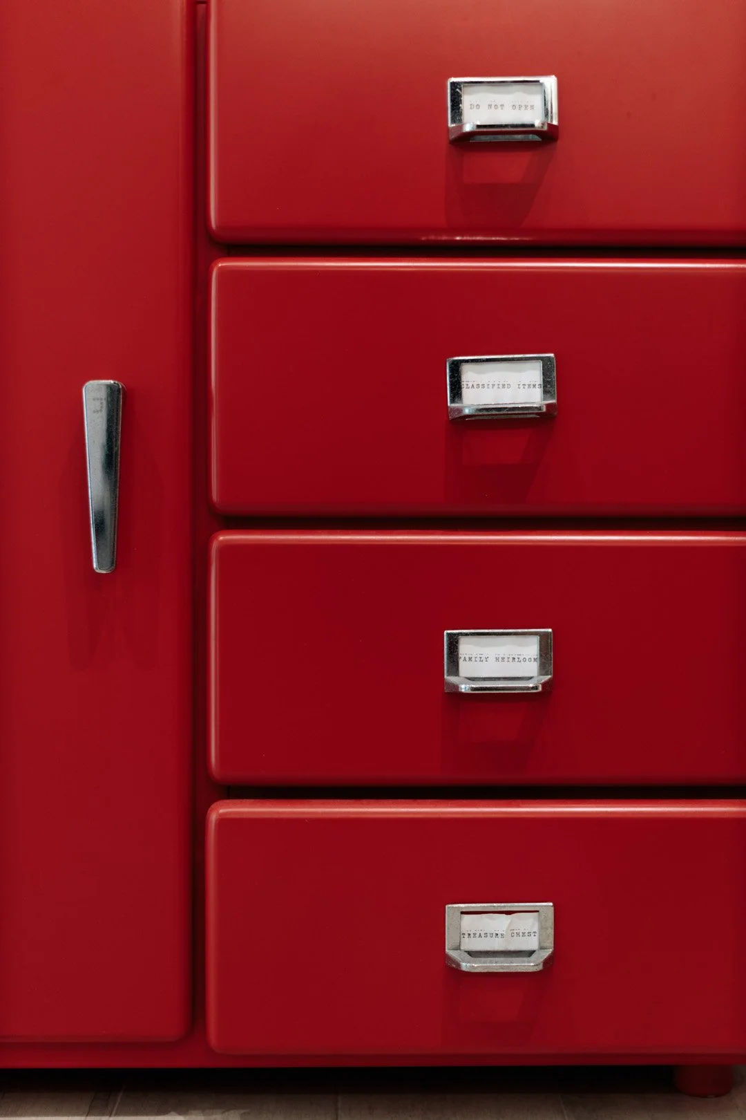 Close-up of a red vintage metal filing cabinet with four drawers, each labeled with vintage typewriter labels. The labels read from top to bottom: "DO NOT OPEN," "CLASSIFIED ITEMS," "FAMILY HEIRLOOM," and "TREASURE CHEST." The cabinet has a silver ha