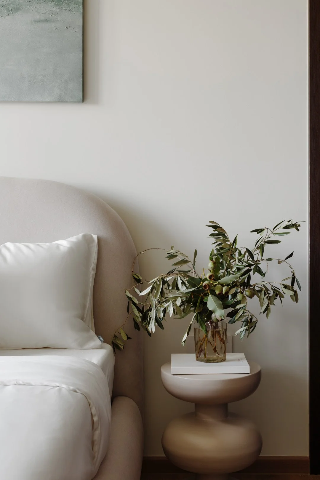 A beige upholstered bed with white bedding and a white pillow, a small round side table with a vase of green leaves and fruit, and a piece of abstract art on the wall in a minimalist bedroom.
