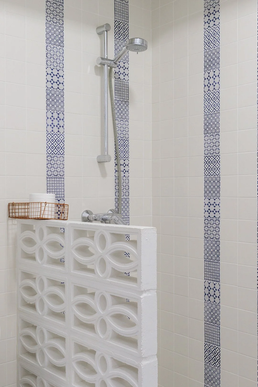 A shower area with white tiled walls decorated with vertical strips of blue patterned tiles. There is a silver showerhead attached to a vertical bar, and a white lattice partition with a decorative pattern separates the shower space. A small copper w