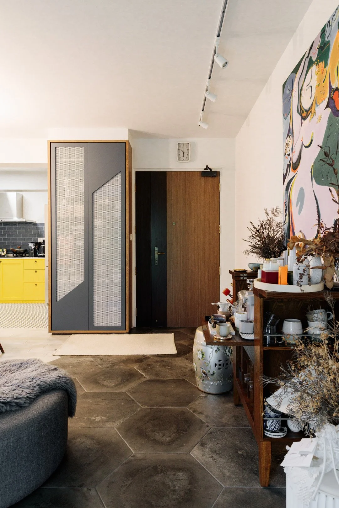 A modern living space with hexagonal stone flooring, a dark wooden door, and a colorful artwork on the wall. There is a wooden console with decorative items and dried plants, and a yellow kitchen visible in the background.