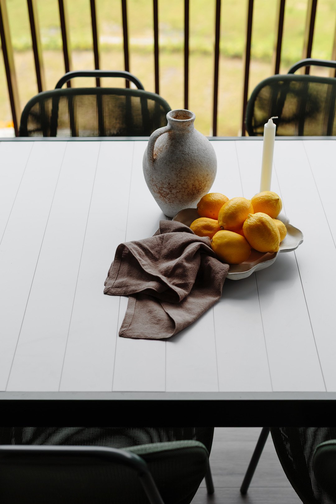A white table with a ceramic vase, a plate of lemons, a single white candle, and a brown cloth, set near a screened porch with black chairs and a view of a grassy yard.