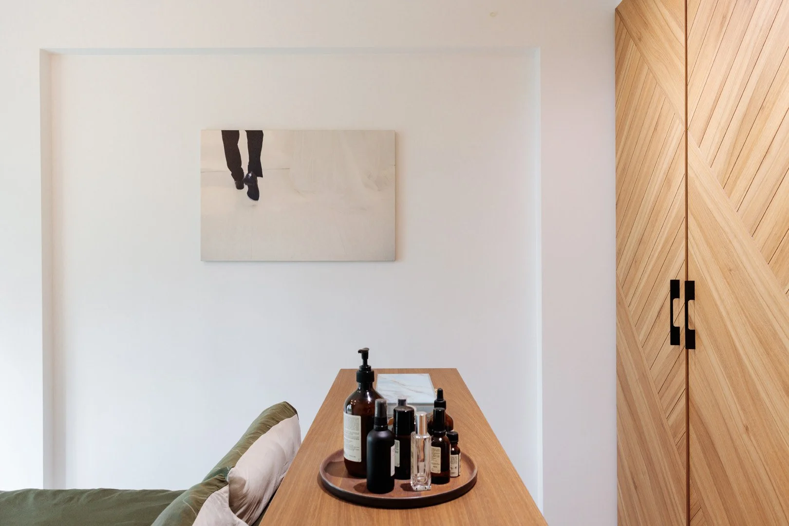 A wooden table with assorted skincare bottles, partly visible green cushion, white wall with abstract black and white artwork and wooden cabinet with chevron pattern.