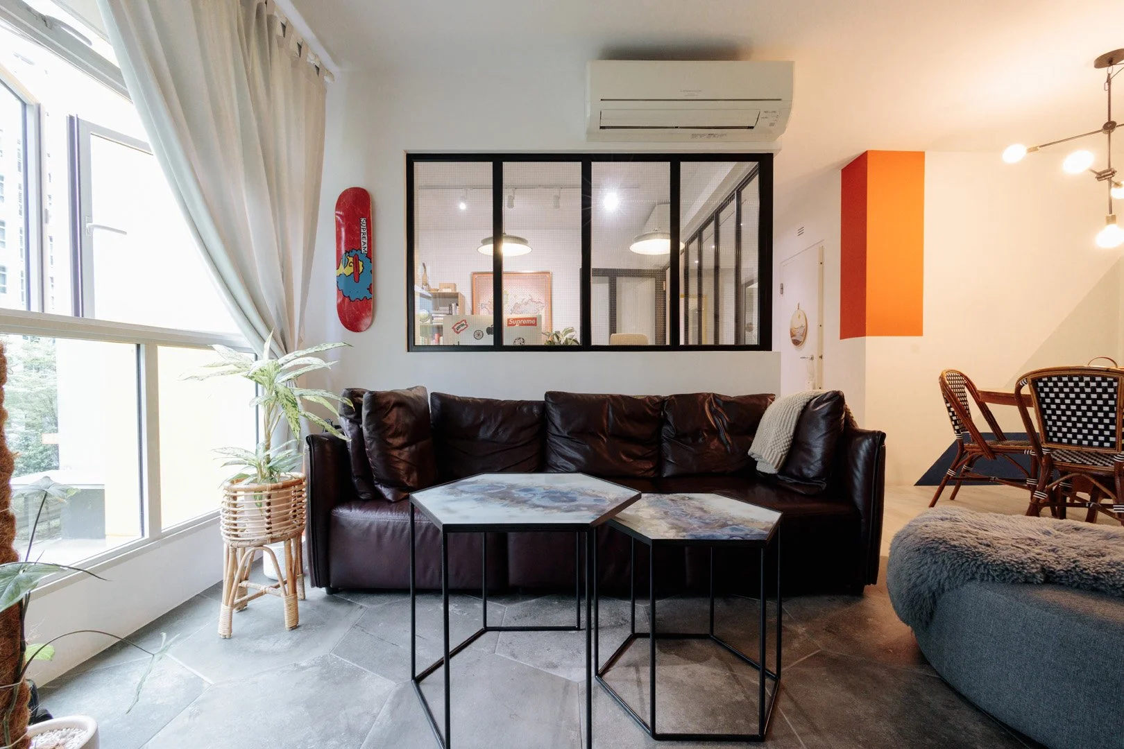 Cozy living room with a brown leather sofa, two matching hexagonal marble-top coffee tables, a large window with white curtains, indoor plants, a skateboard wall decoration, and a glass partition separating the living space from a work area.