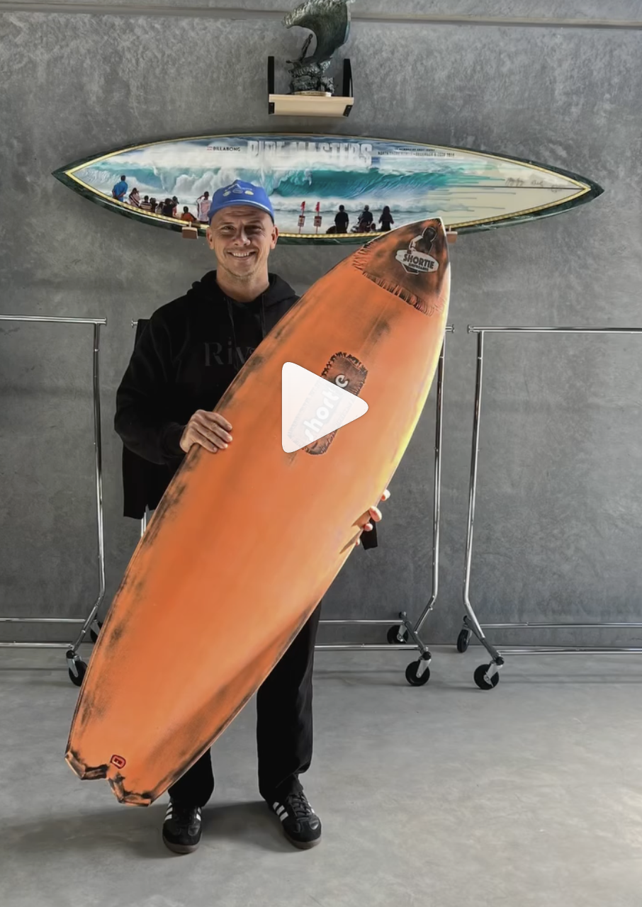 Julian Wilson, standing with a custom Shortie Surfboard in his warehouse in front of his Pipe masters trophy.  See Julian Wilson surf on a custom Shortie Surfboard.