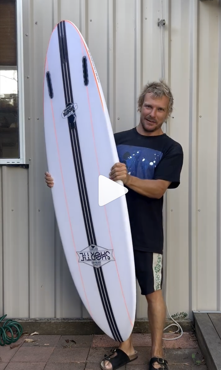 Sam Ayton, Shortie Surfboard shaper, explaining his white surfboard model with a carbon fibre stringer, standing outside near a metal siding wall.