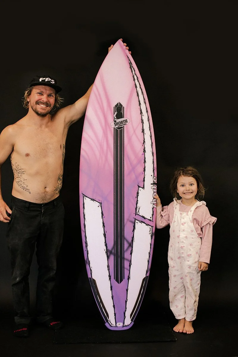 Sam Ayton, Shortie Surfboard custom surfboard shaper, standing with his popular Crazy Swayze model all-rounder surfboard - best ridden in all conditions - painted pink and purple, with his daughter holding the other side of the surfboard.