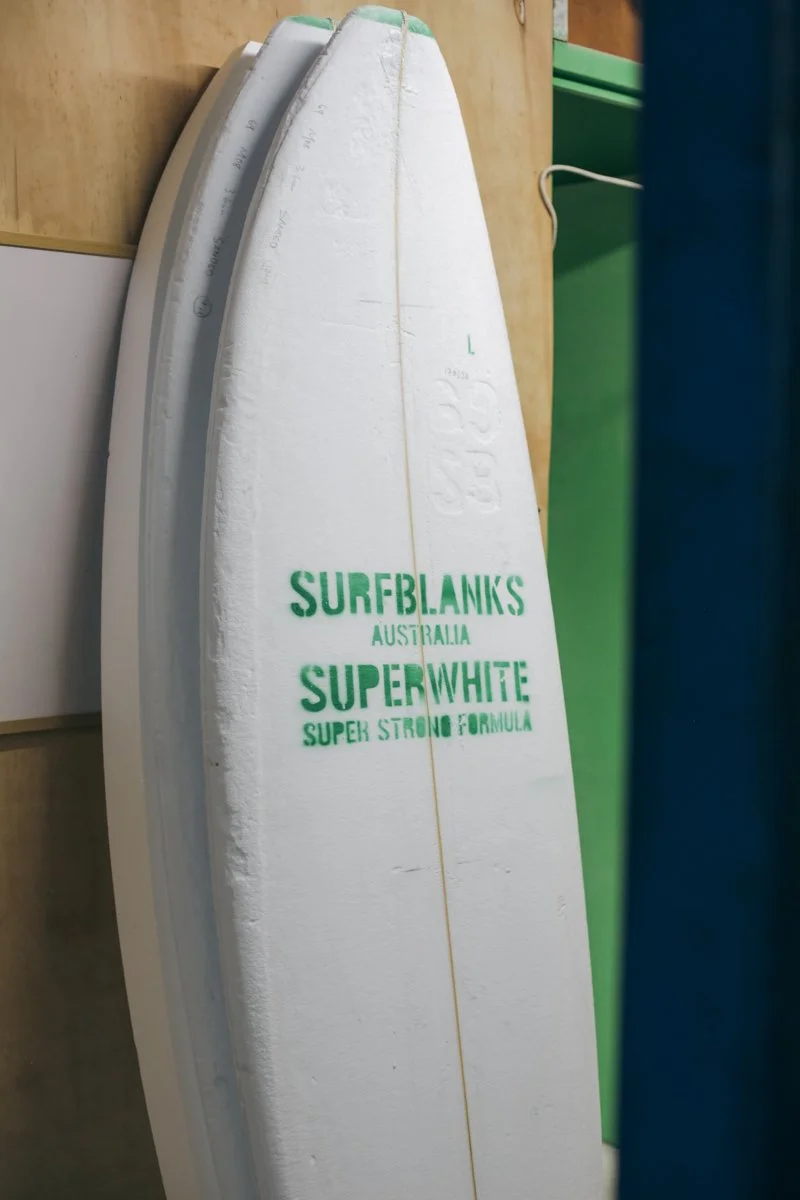 A surfboard with green text reading 'SURFBLANKS AUSTRALIA SUPERWHITE SUPER STRONG FORMULA' leaning against a wooden surface.