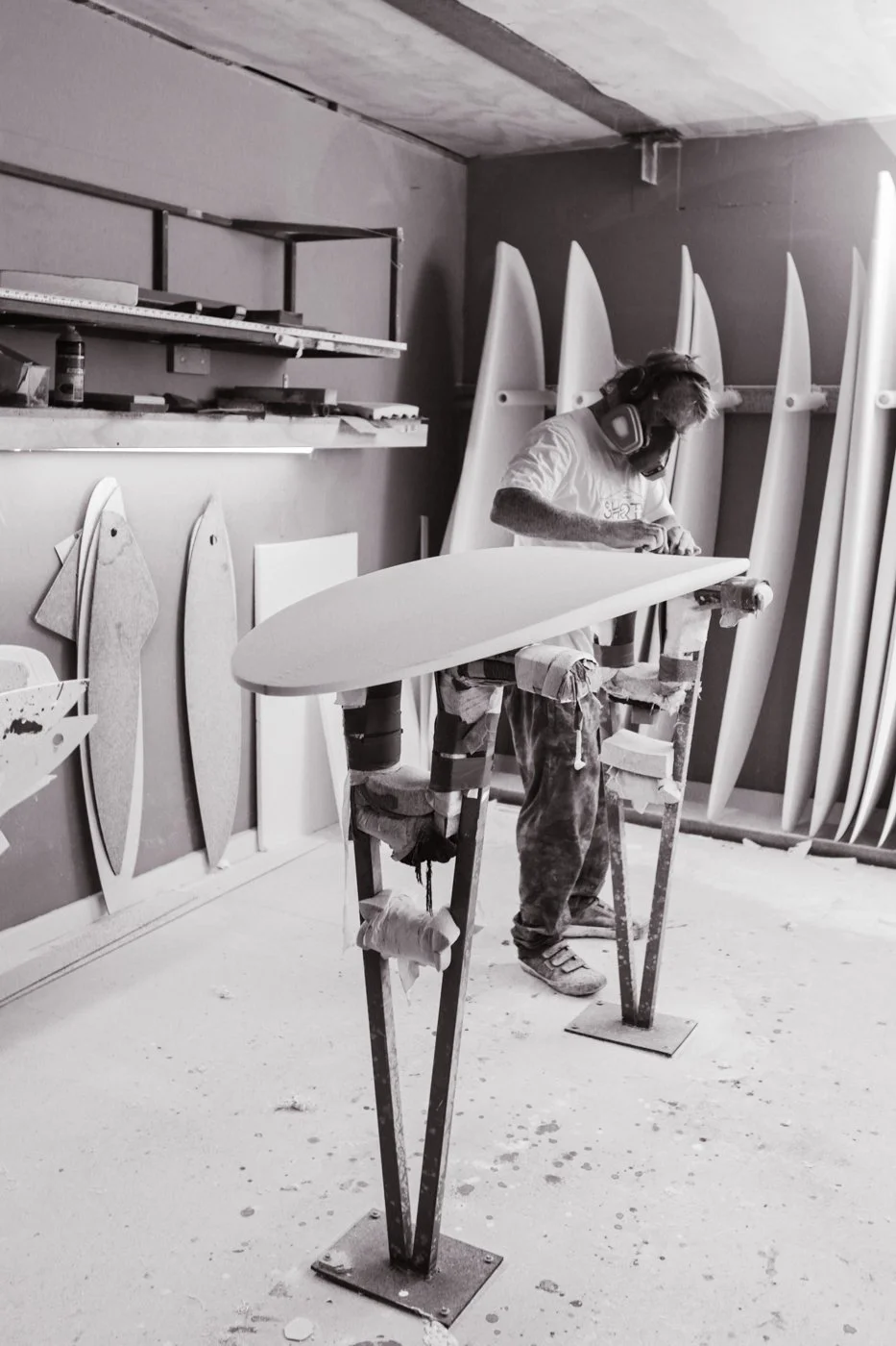 A person working on a surfboard in a workshop, wearing headphones and safety glasses, with multiple surfboards leaning against the wall in the background.