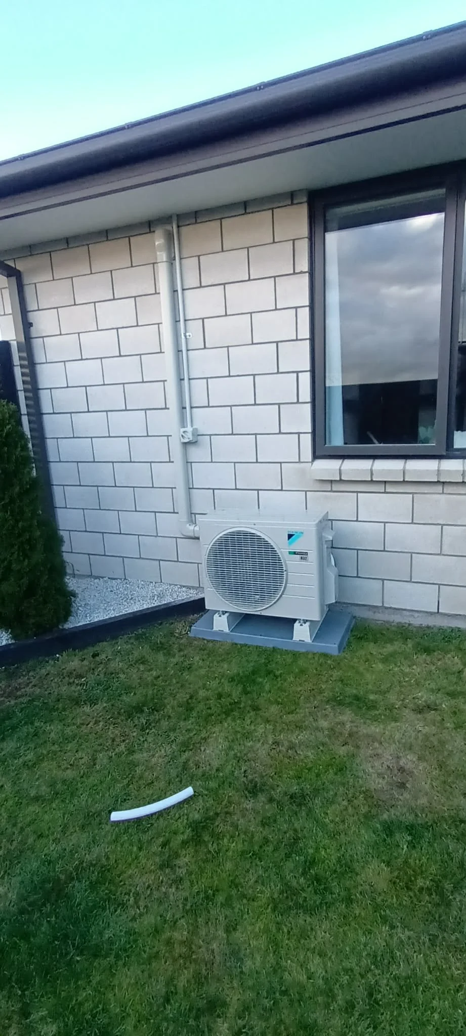 Outside view of a house wall with an air conditioning unit installed on the ground, a window, house's brick wall, and a small bush on the left side.