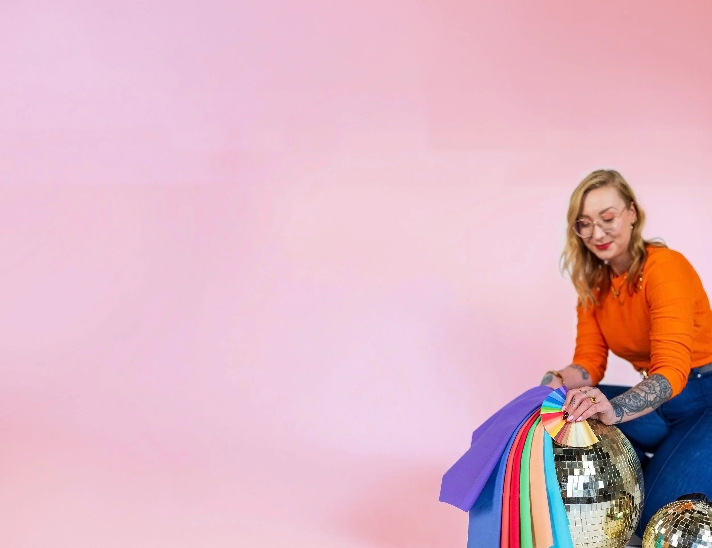 A woman with glasses, orange top, and tattoos on her arms, sitting against a pink background, holding a fan with numerous color swatches.