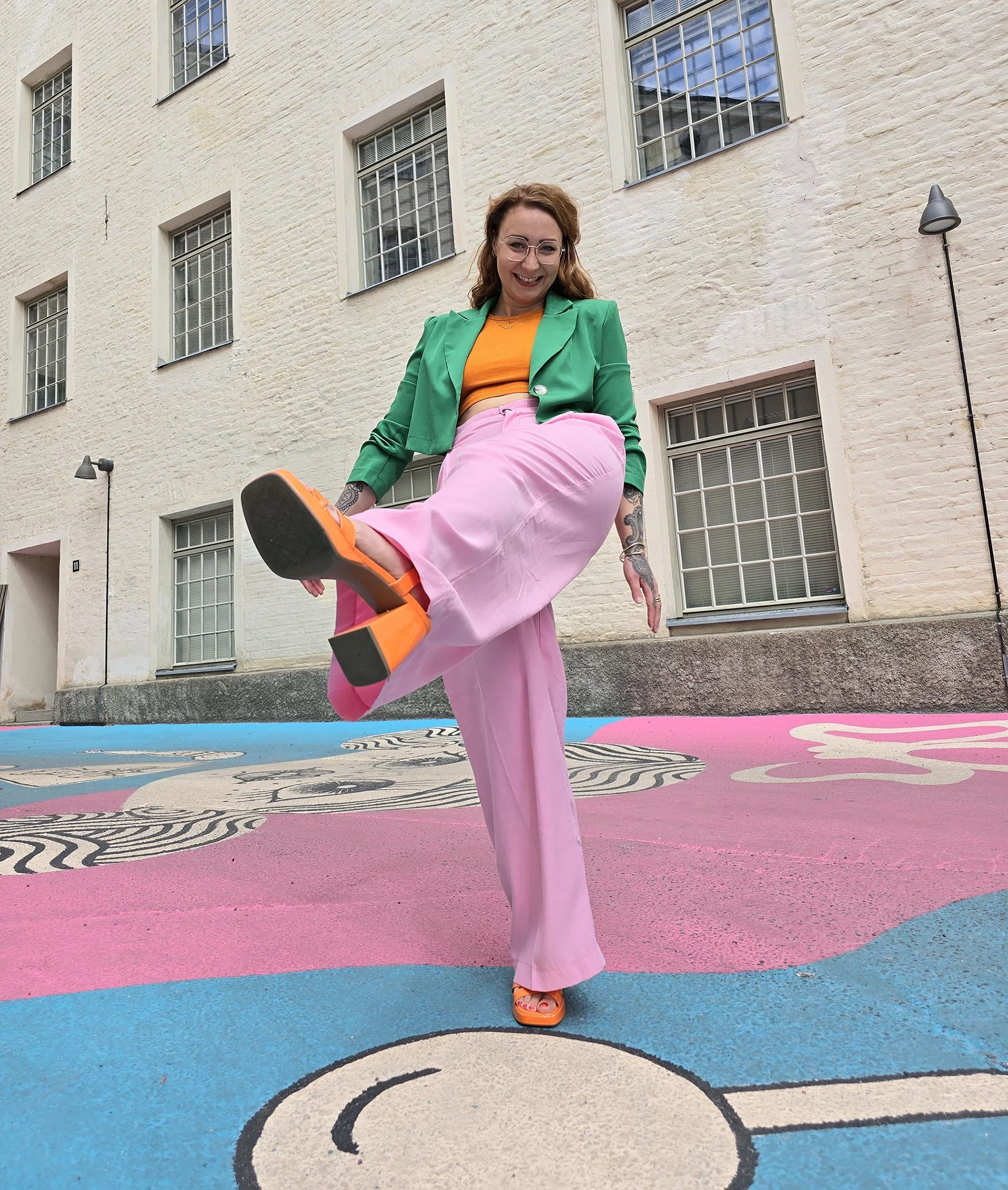 A woman wearing a green blazer, orange top, pink pants, and orange platform shoes, smiling and kicking her foot toward the camera. She has glasses and tattoos on her arms. She is outdoors on a colorful painted surface with a white brick building in the background.