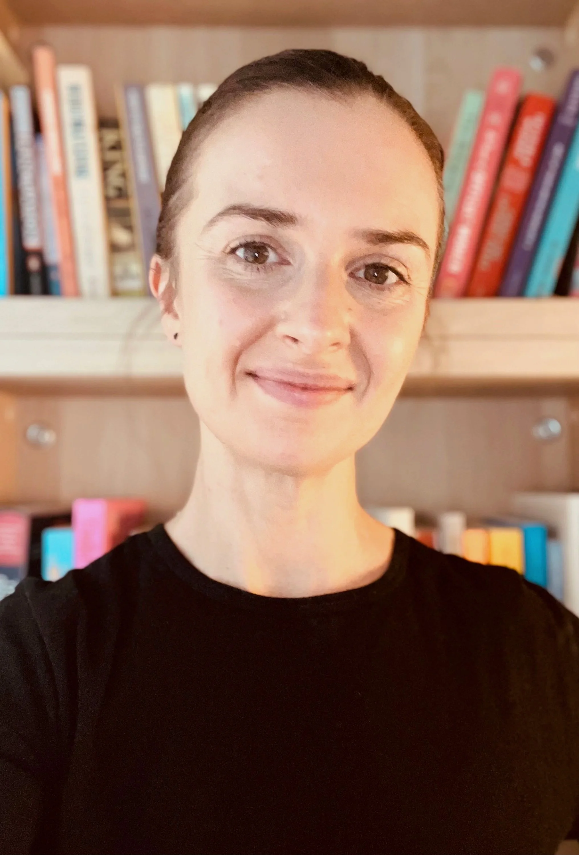 A woman with short dark hair and a black shirt smiling, standing in front of a bookshelf filled with colorful books.