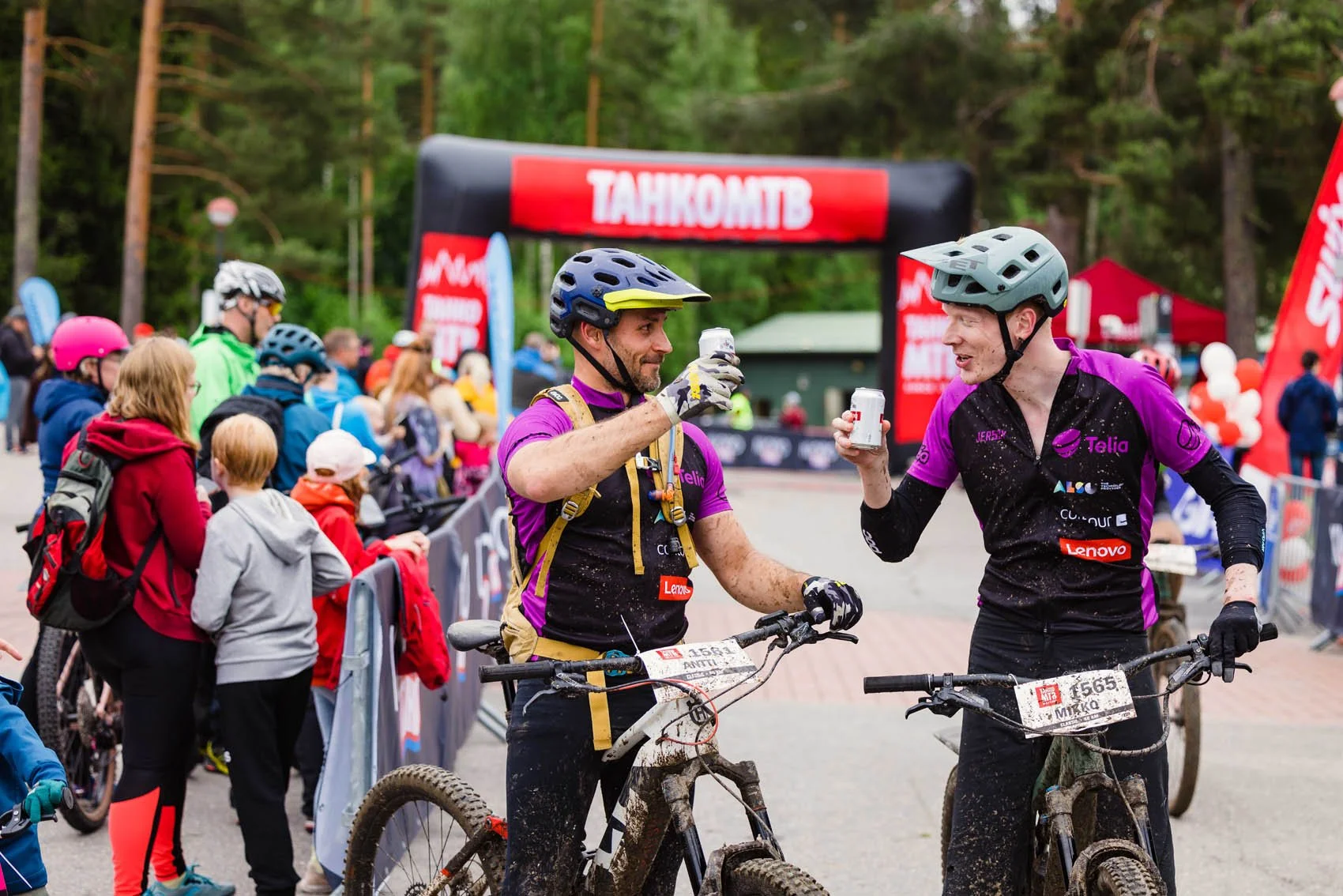 Kaksi pyöräilijää maalialueella juoden yhdessä urheilujuomaa, pyörät likaantuneita ja ihmisjoukko katsomassa taustalla.