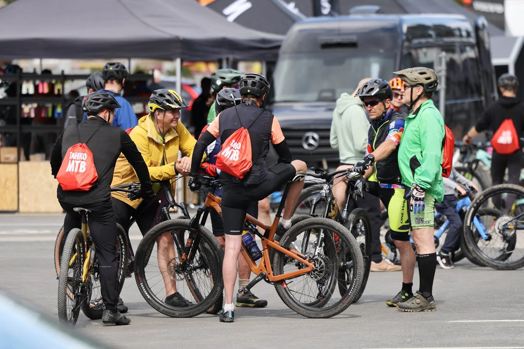 Ryhmä ihmisiä keskustelee keskenään. Heillä on pyörät, kypärät ja urheiluasut, ja taustalla on telttoja ja muita ihmisiä.