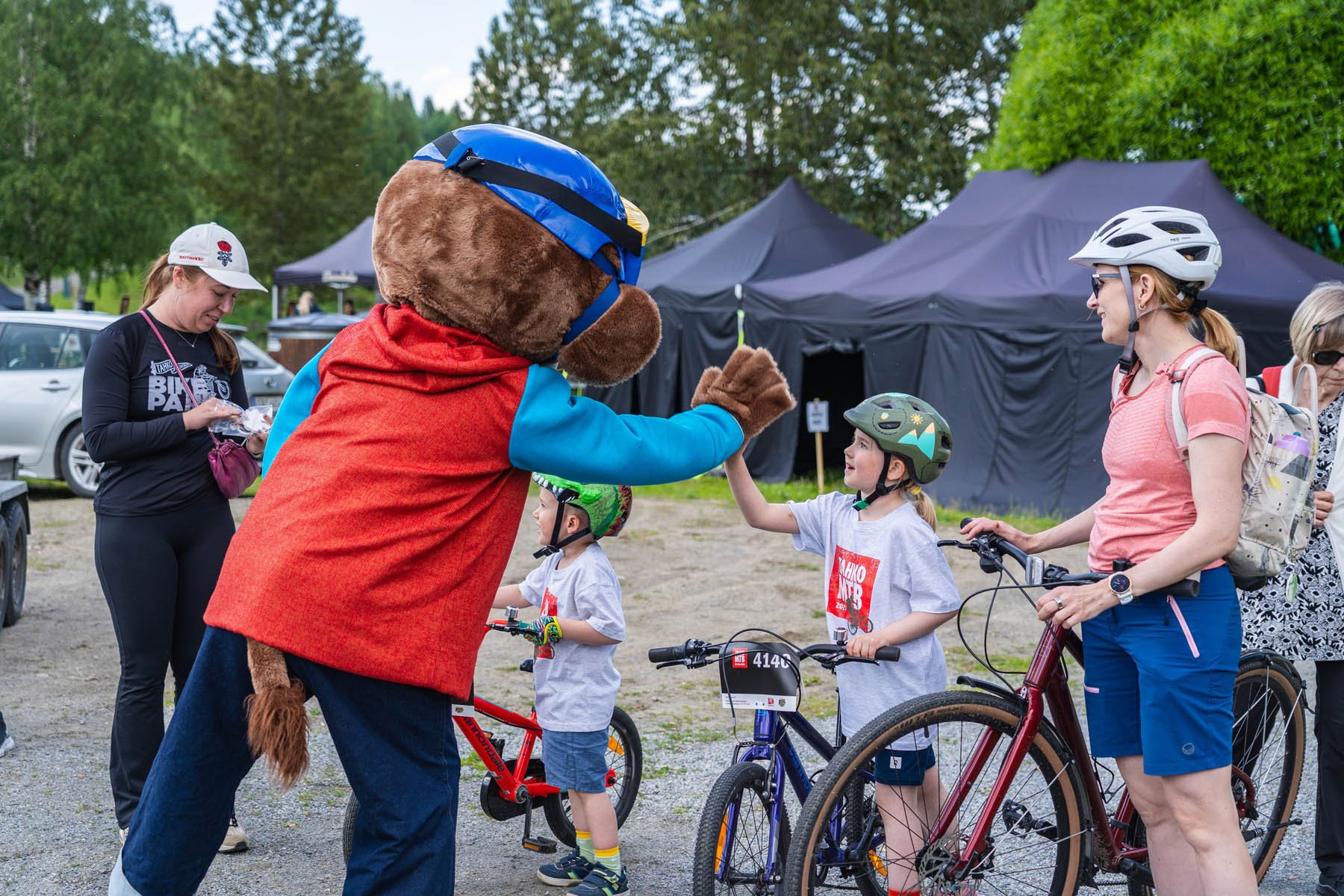 Ihminen on pukeutunut maskotiksi ja hän juttelee lapsille ja aikuisille, jotka ovat osallistumassa pyöräkilpailuun.