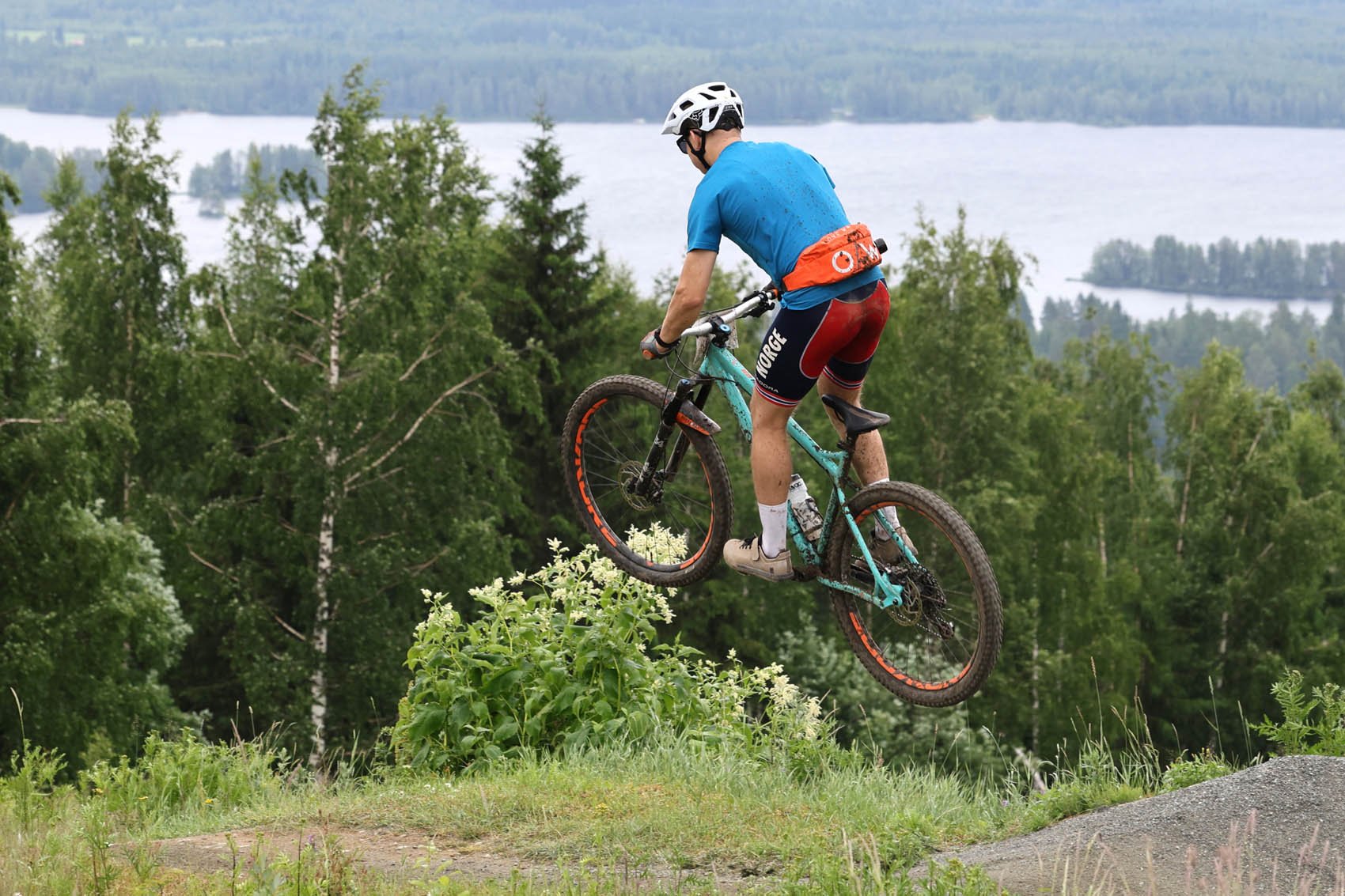 Mies pyöräilee polkupyörällä korkealla mäellä, taustalla järvi ja metsää.