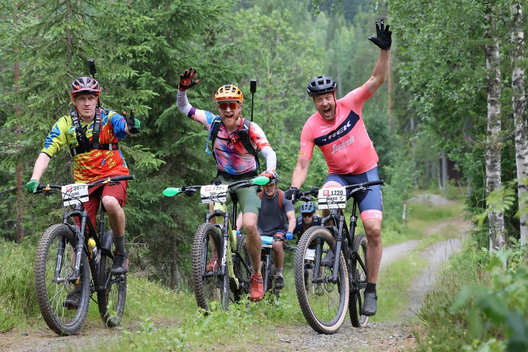 Kolme pyöräilijää metsässä, kaikki ovat iloisia ja he nostavat kättä yleisölle, pyörissä on numerolaput.