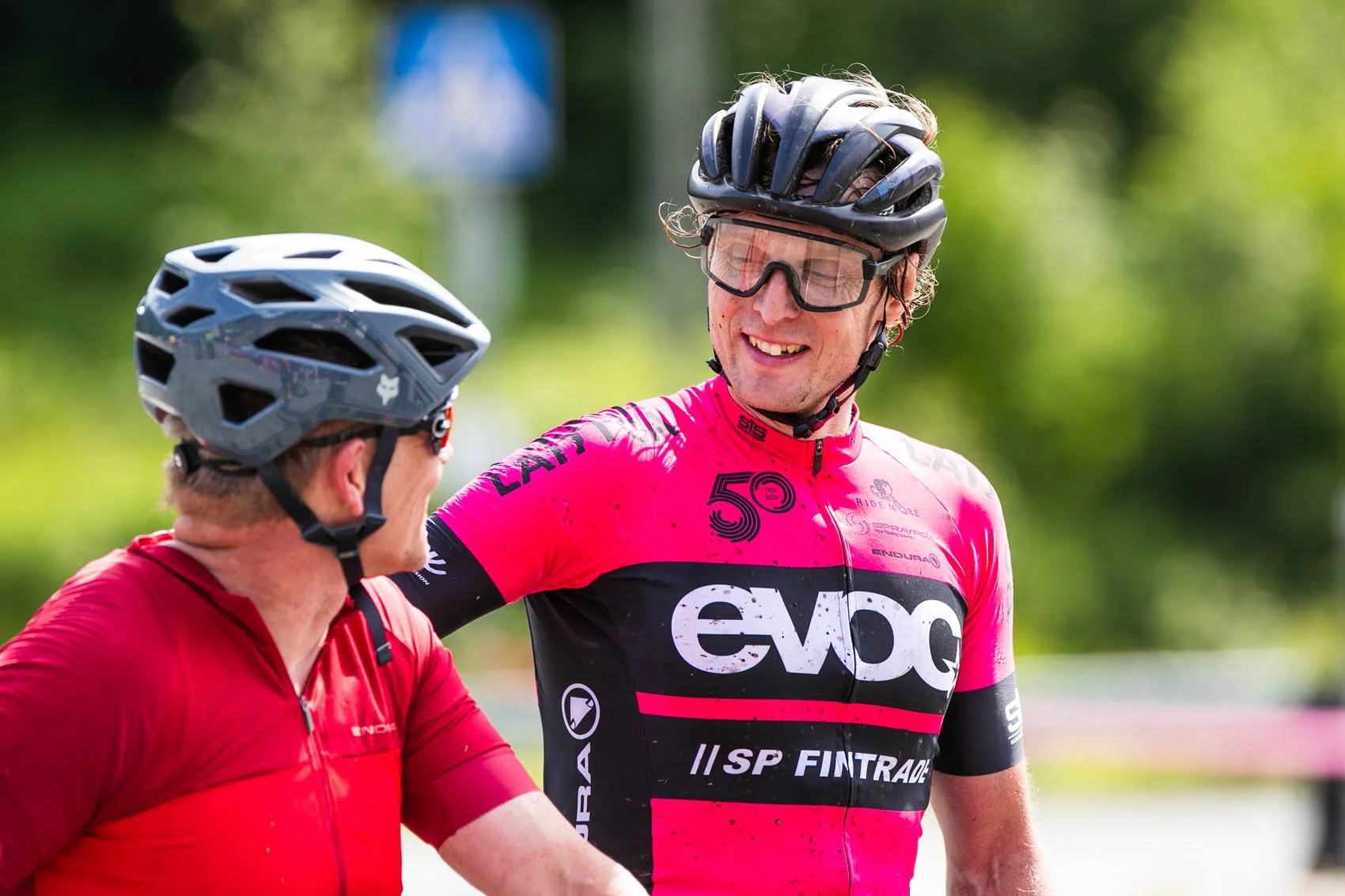 Kaksi pyöräilijää hymyilee ja keskustelee, heillä on kypärät ja ajolasit, etualalla yksi pyöräilijä ja takana toinen, taustalla vihreä luonto.
