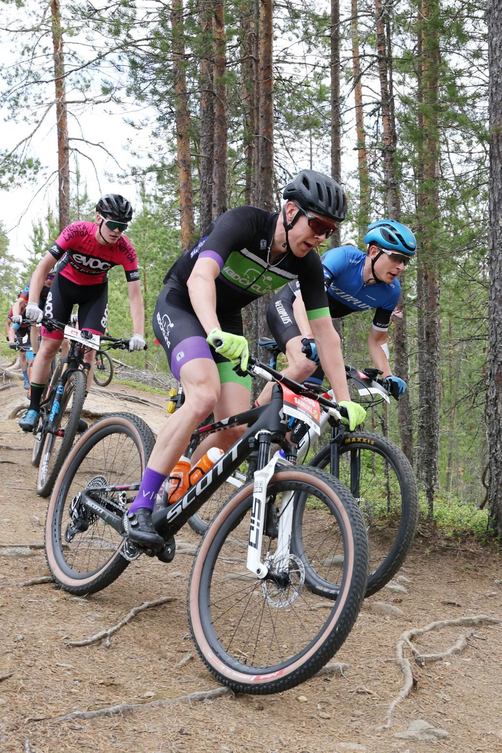 Kolme pyöräilijää ajaa kovaa vauhtia maastopyörillä metsässä.