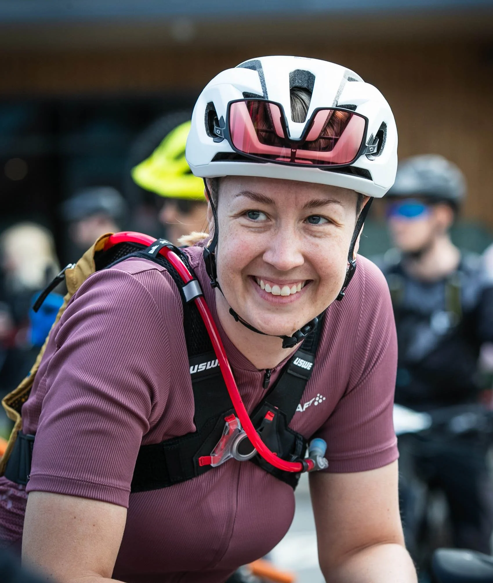 Nainen hymyilee. Hänellä on pyöräilykypärä päässään ja taustalla on muita pyöräilijöitä.