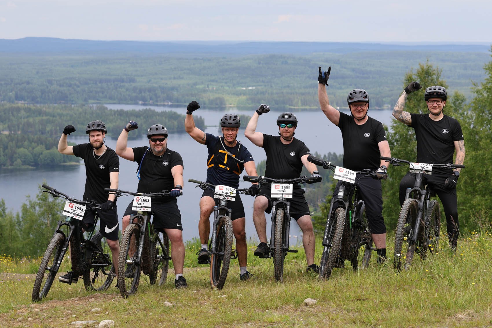 Seitsemän miestä seiso polkupyörien kanssa, kaikilla kypärät ja mustat urheilupaidat, taustalla järvi ja vihreitä puita, aurinkoinen sää.