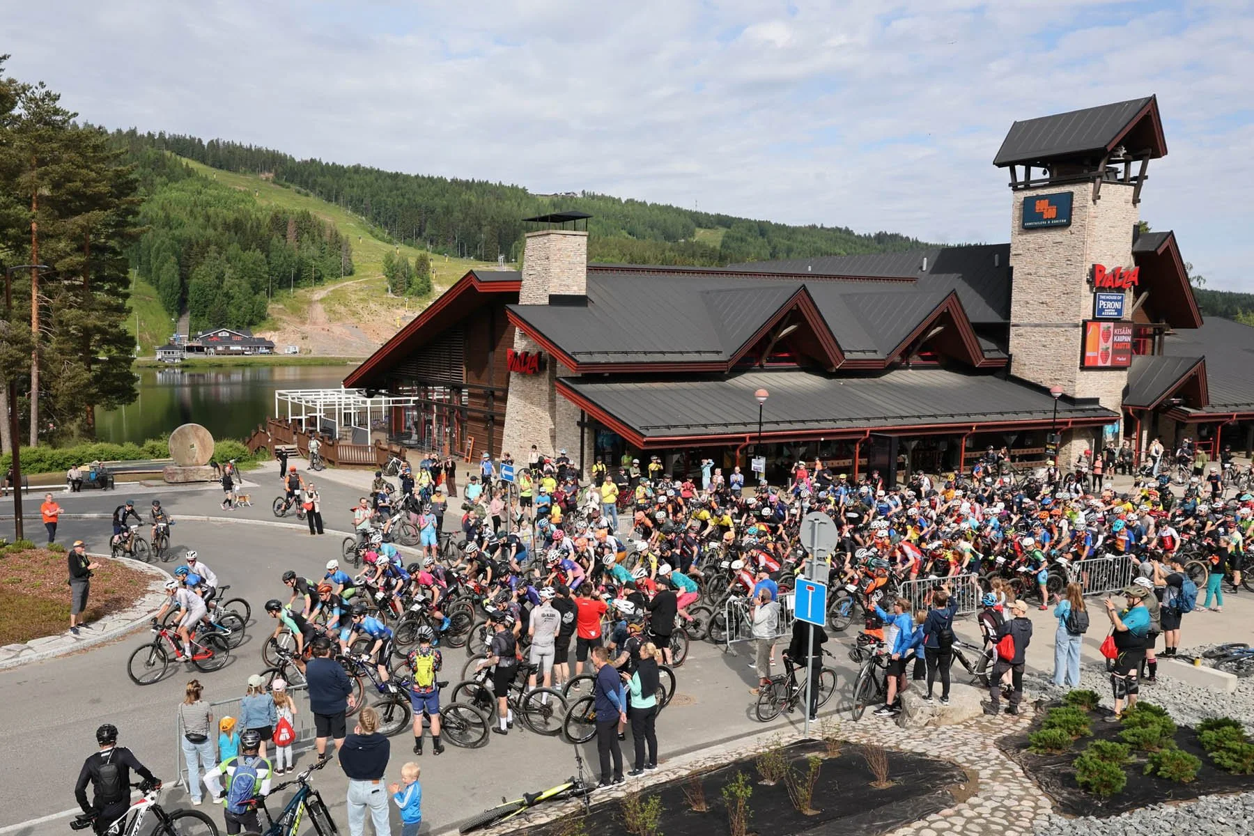Ihmiset ovat kerääntyneet aukiolle katsomaan pyöräkilpailun lähtöä. Taustalla on rakennus veden rannalla.