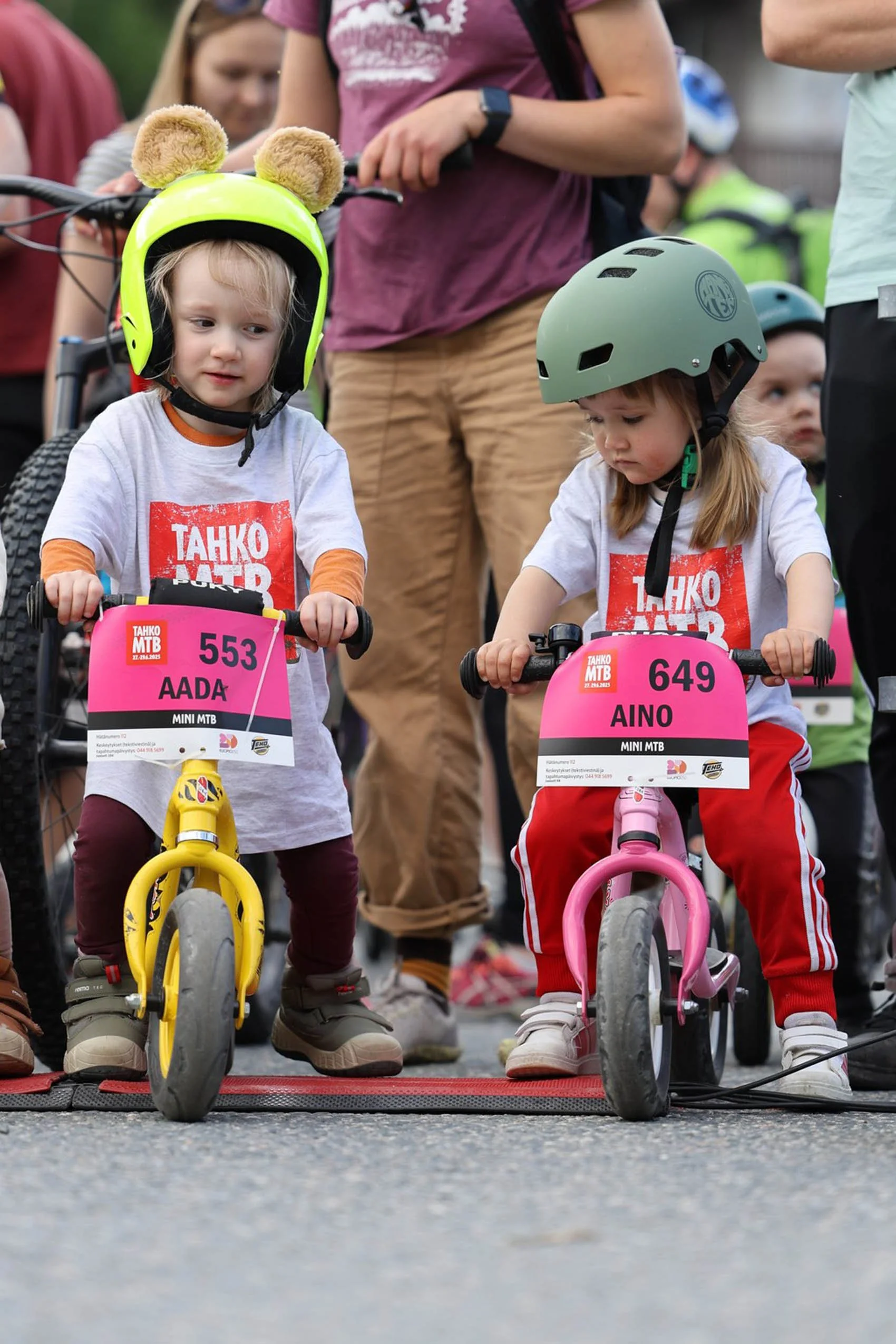 Kaksi pientä lasta pyöräkilapailun starttialueella, toisella keltainen pyörä ja toinen pinkki, molemmilla on kypärät ja osallistumisnumerot paidassa. Ympärillä aikuisia ja muita lapsia.