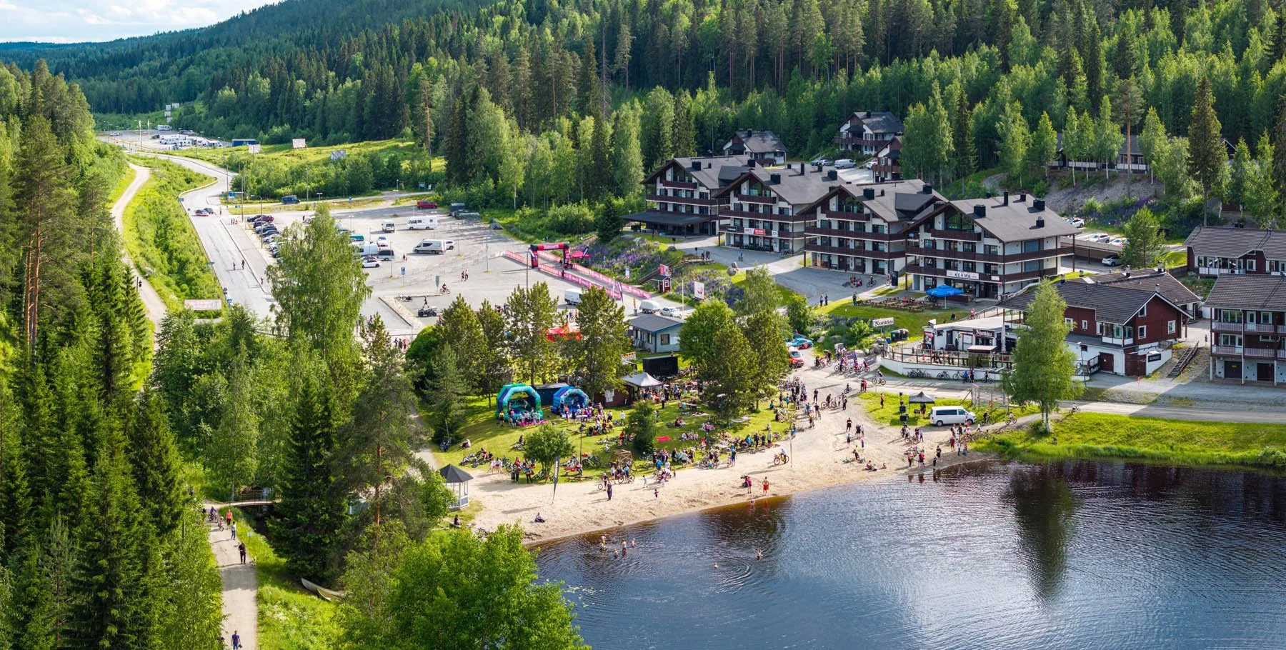 Ilmakuva järven rannasta, jossa on ihmisiä nauttimassa auringosta. Taustalla näkyy majoitusrakennuksia ja mäkiä.