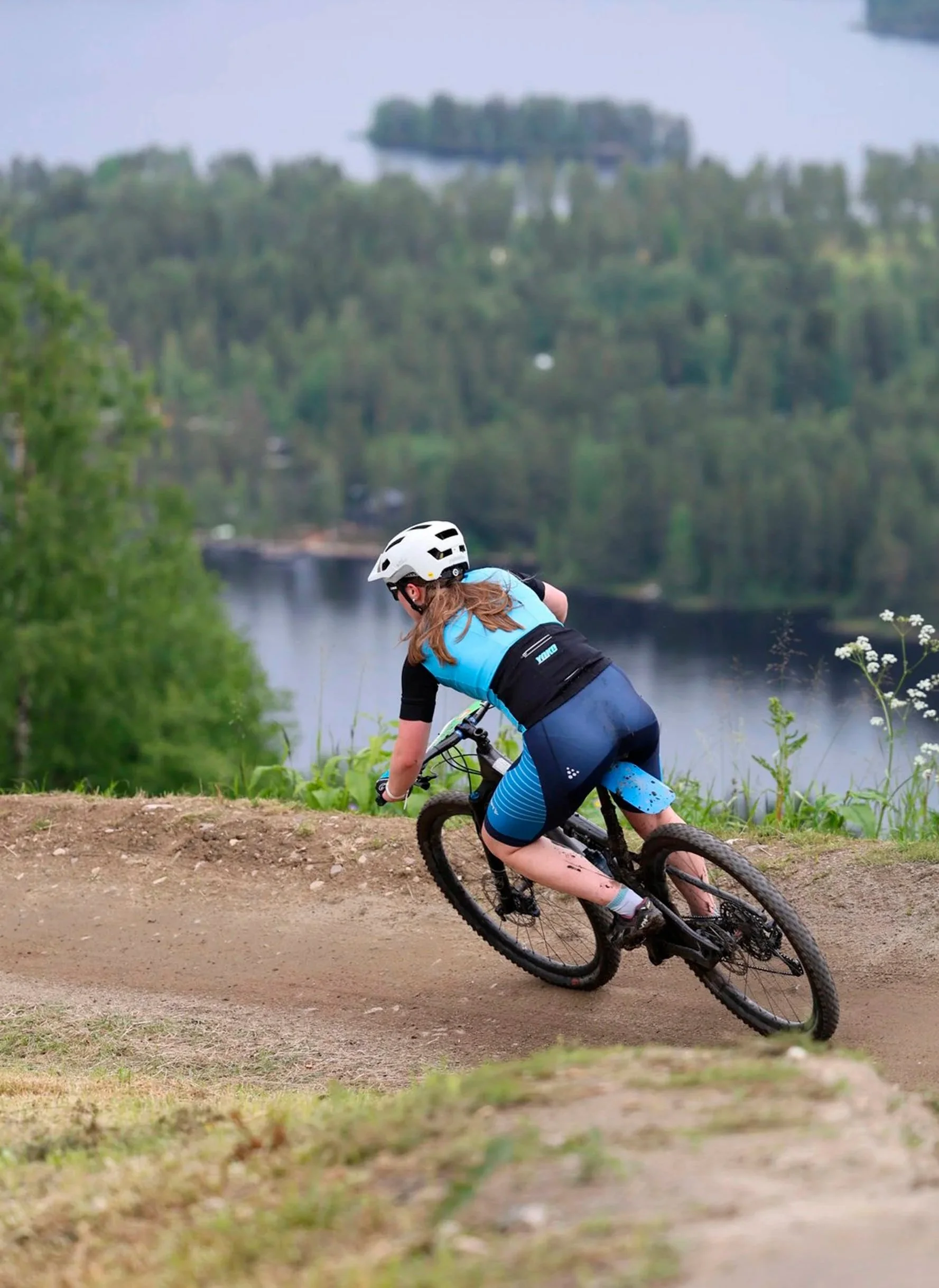 Kilpailija pyöräilee alamäkeä metsäisellä polulla, taustalla joki ja puita.