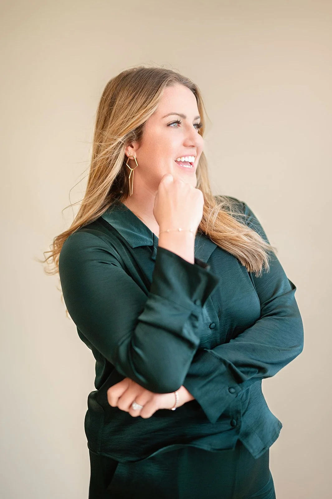 A woman with long blonde hair wearing a dark green shirt and gold earrings, smiling and looking to the side, with her hand near her face.