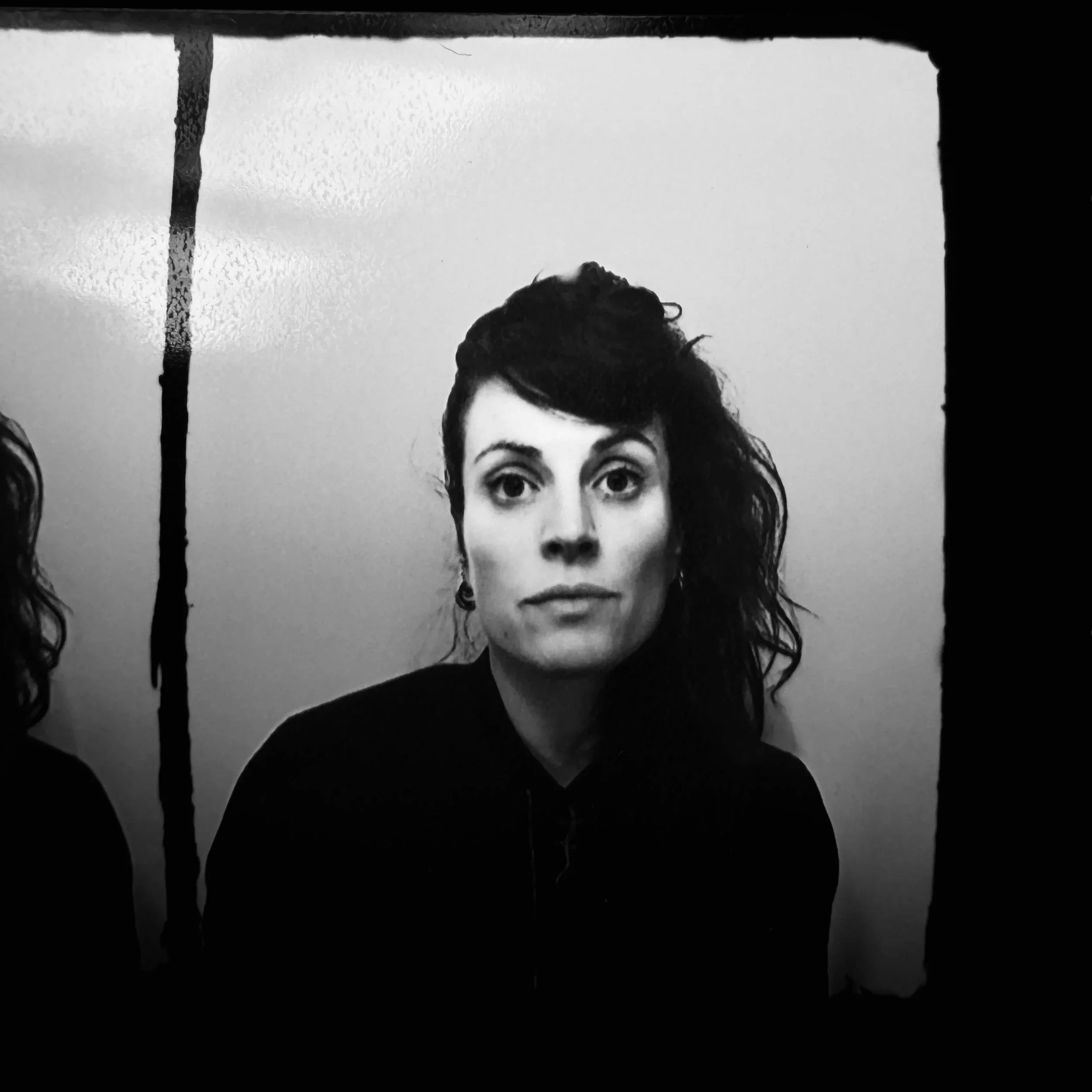 Portrait en noir et blanc de Sandrine Lopez avec cheveux foncés et ondulés, portant un haut foncé, regardant directement l'objectif, avec un fond clair.