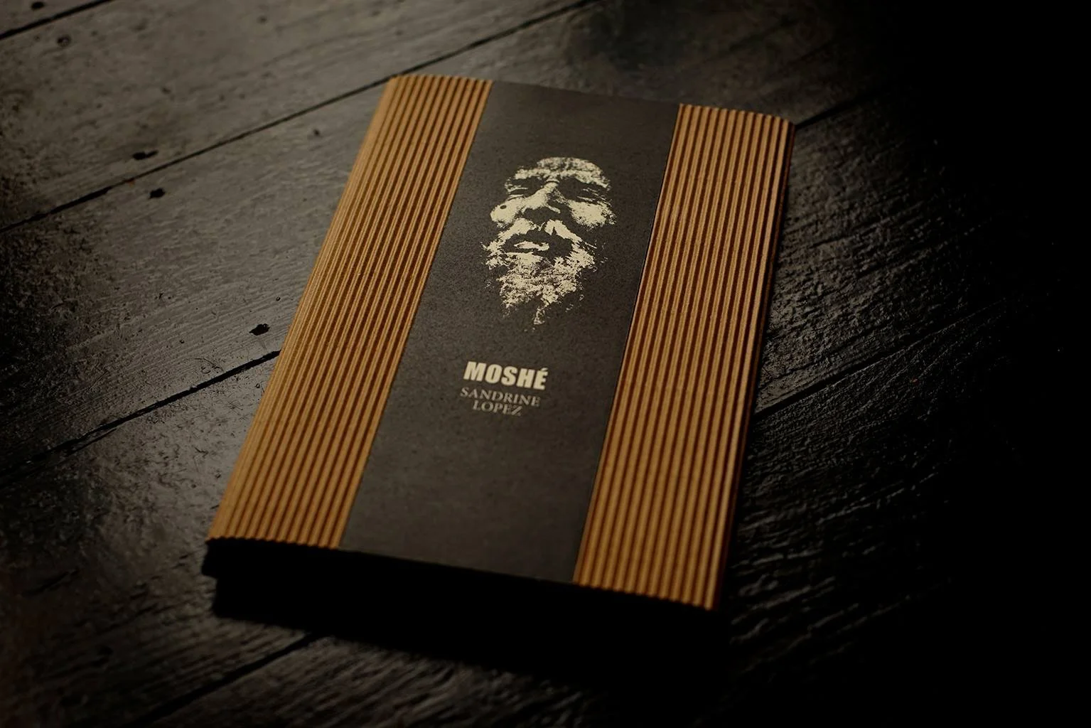 Livre avec une couverture noire et des rayures en bois, portant le titre 'MOSHÉ' et le nom 'SANDRINE LÓPEZ', avec une illustration blanche de face d'une personne âgée, probablement Moshe.