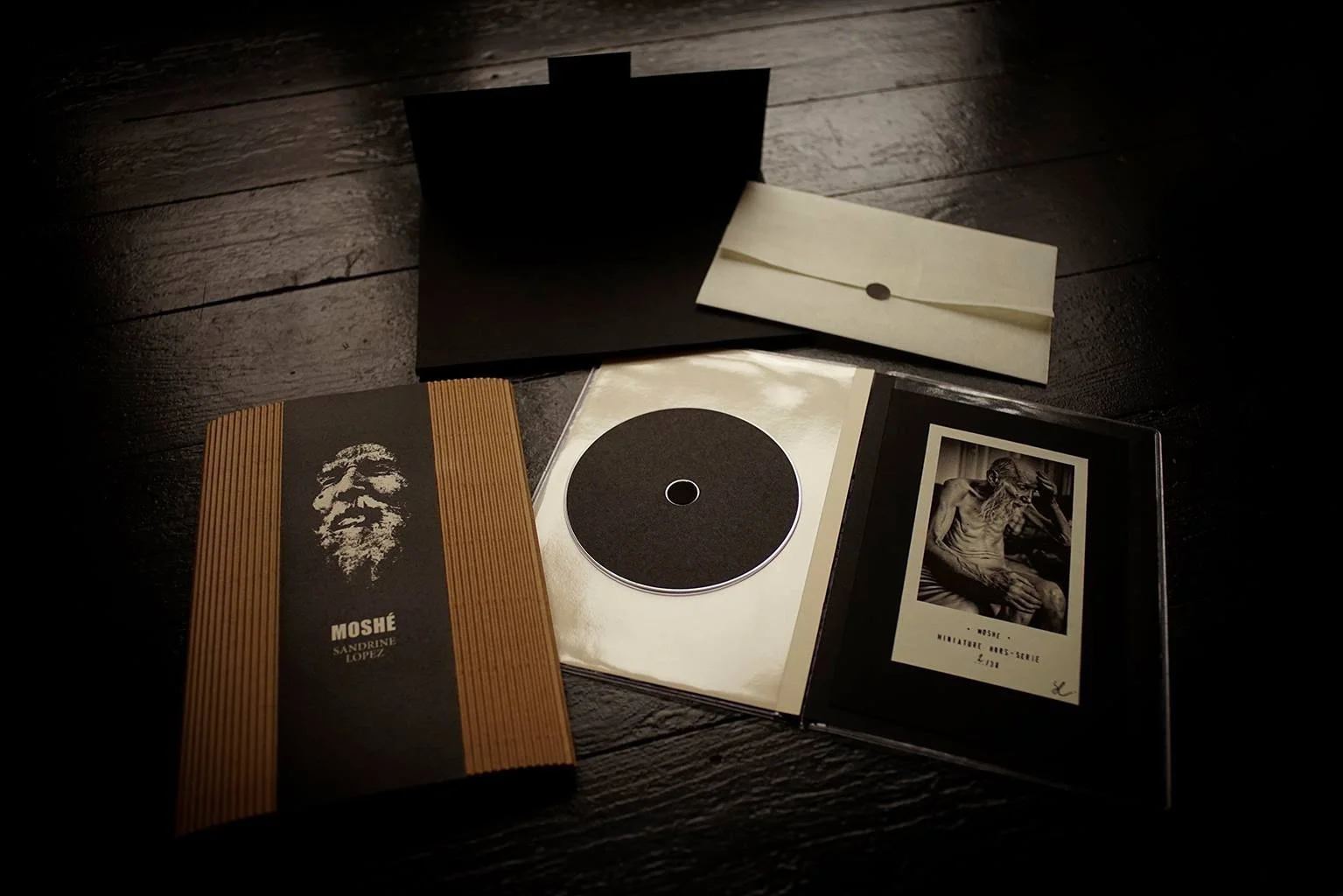 Collection de livres et de documents liés à une personne nommée Moshe, comprenant un livre avec une illustration d'un visage barbu, un CD ou DVD, et une photo en noir et blanc d'un homme âgé assis, tous disposés sur un plan en bois sombre.