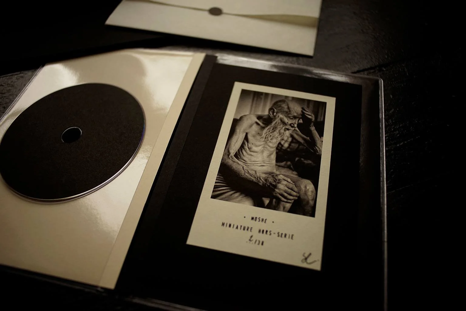 Photo en noir et blanc d'une vieille statue d'homme, assis, avec une barbe longue, une main sur la tête et une autre sur le genou, avec des livres en arrière-plan. Planches de papier et un cd sont visibles à côté.