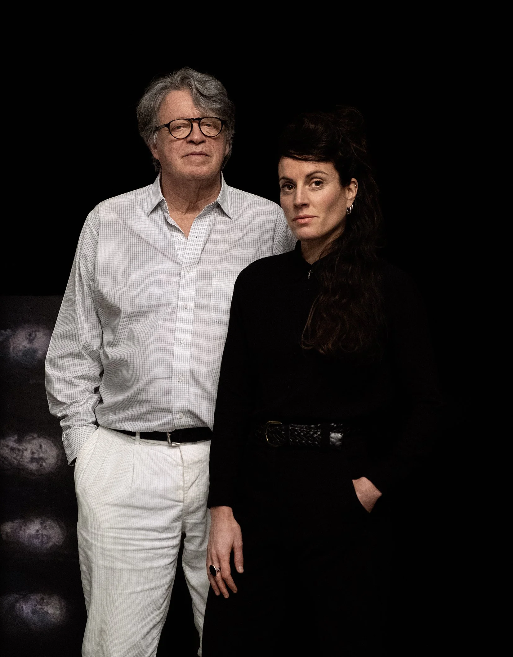 Un homme et une femme posent ensemble devant un fond noir. L'homme porte une chemise claire à manches longues et un pantalon blanc, il a des lunettes et des cheveux gris. La femme porte un vêtement noir, a de longs cheveux bruns foncés et un maquillage léger.