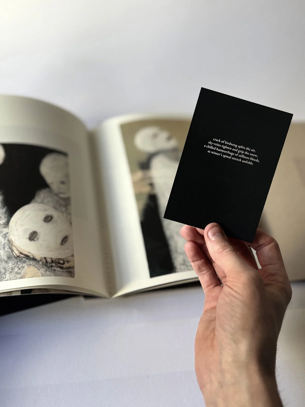 Une main tient une petite carte noire avec un texte poétique blanc. Un livre ouvert avec des images en noir et blanc de personnes est posé en arrière-plan sur une surface blanche.