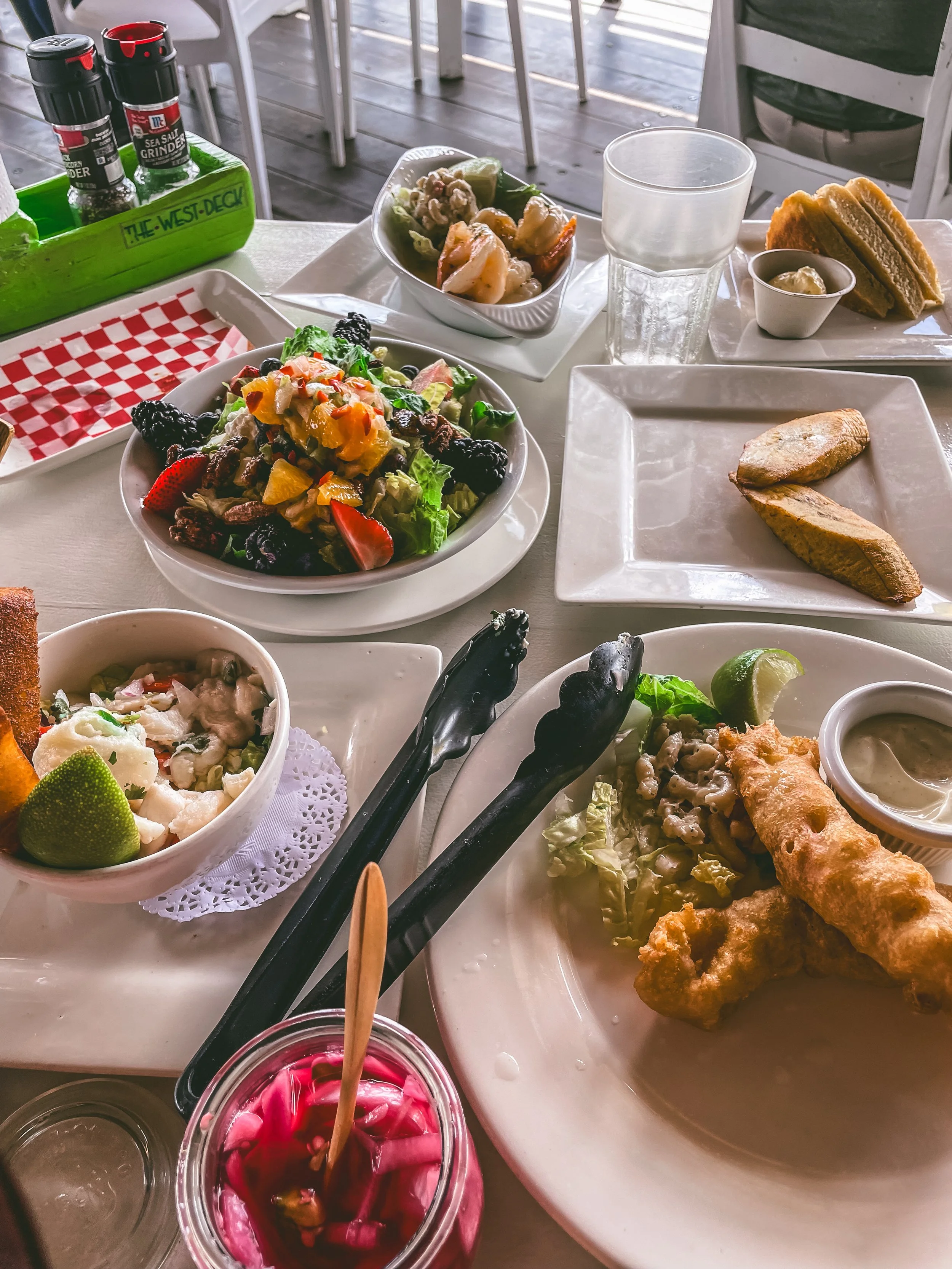 A table of assorted foods including a colorful salad, grilled chicken, fried fish, sandwiches, and condiments on a dining table.