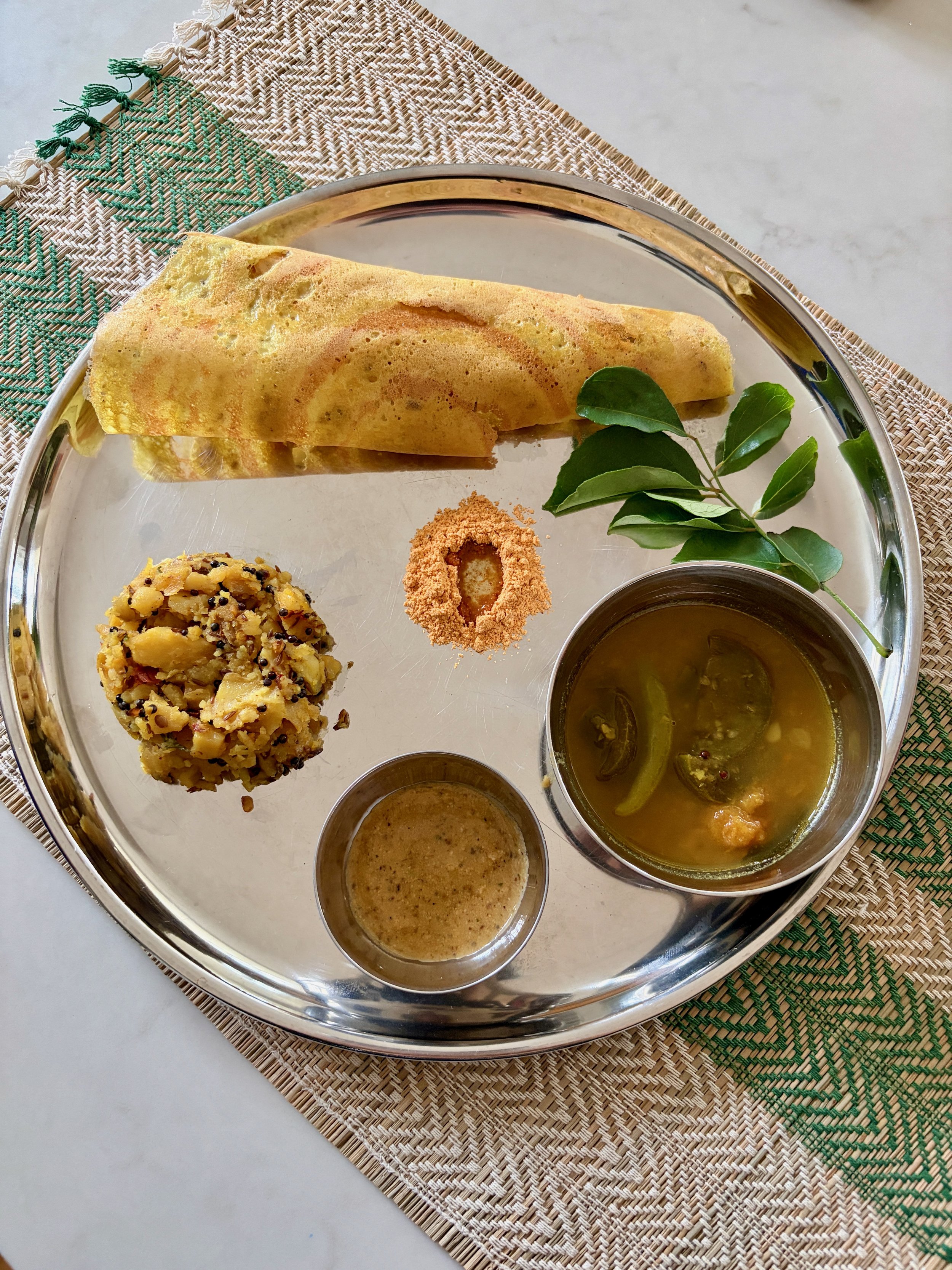 A traditional Indian vegetarian meal served on a round stainless steel tray. The meal includes a dosa, a type of thin crepe, along with a ball of spicy mashed potatoes, a bowl of vegetable sambar, and a small cup of coffee or tea. The tray is placed on a woven placemat.