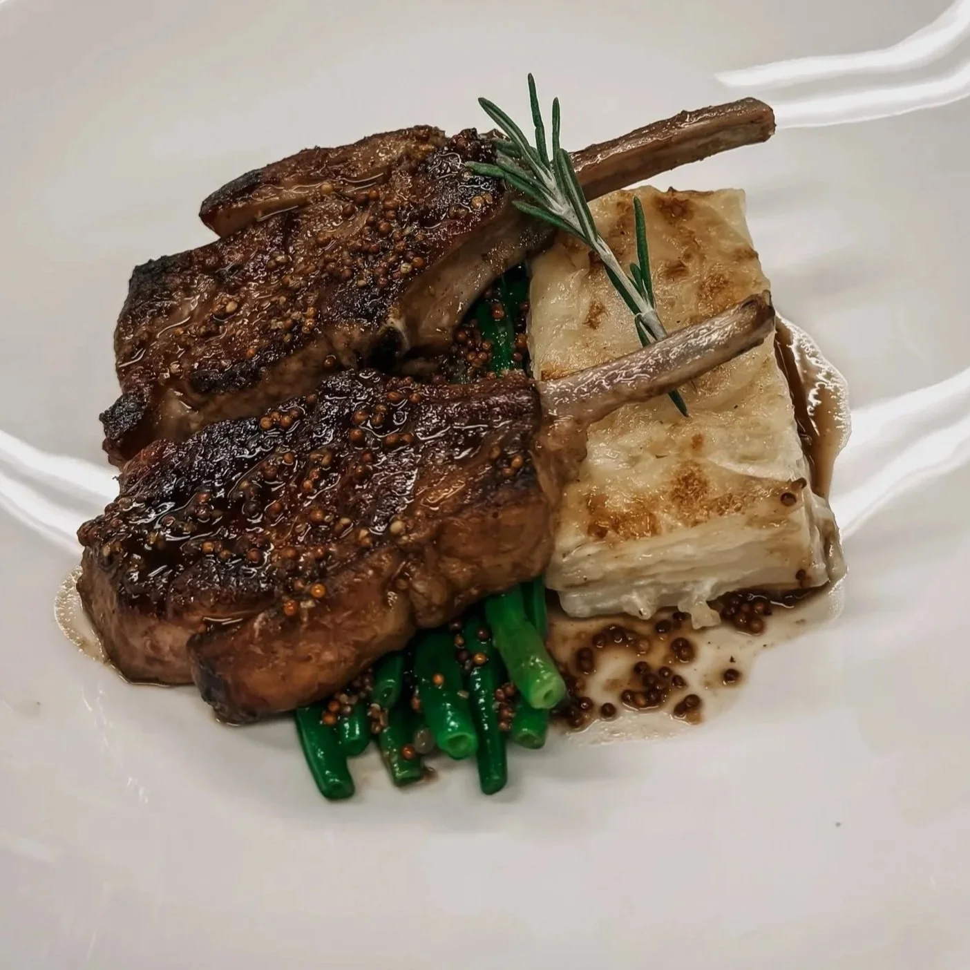 Pan-seared rack of lamb with a sprig of rosemary, served with green beans and potato gratin, topped with mustard seeds and a light sauce.