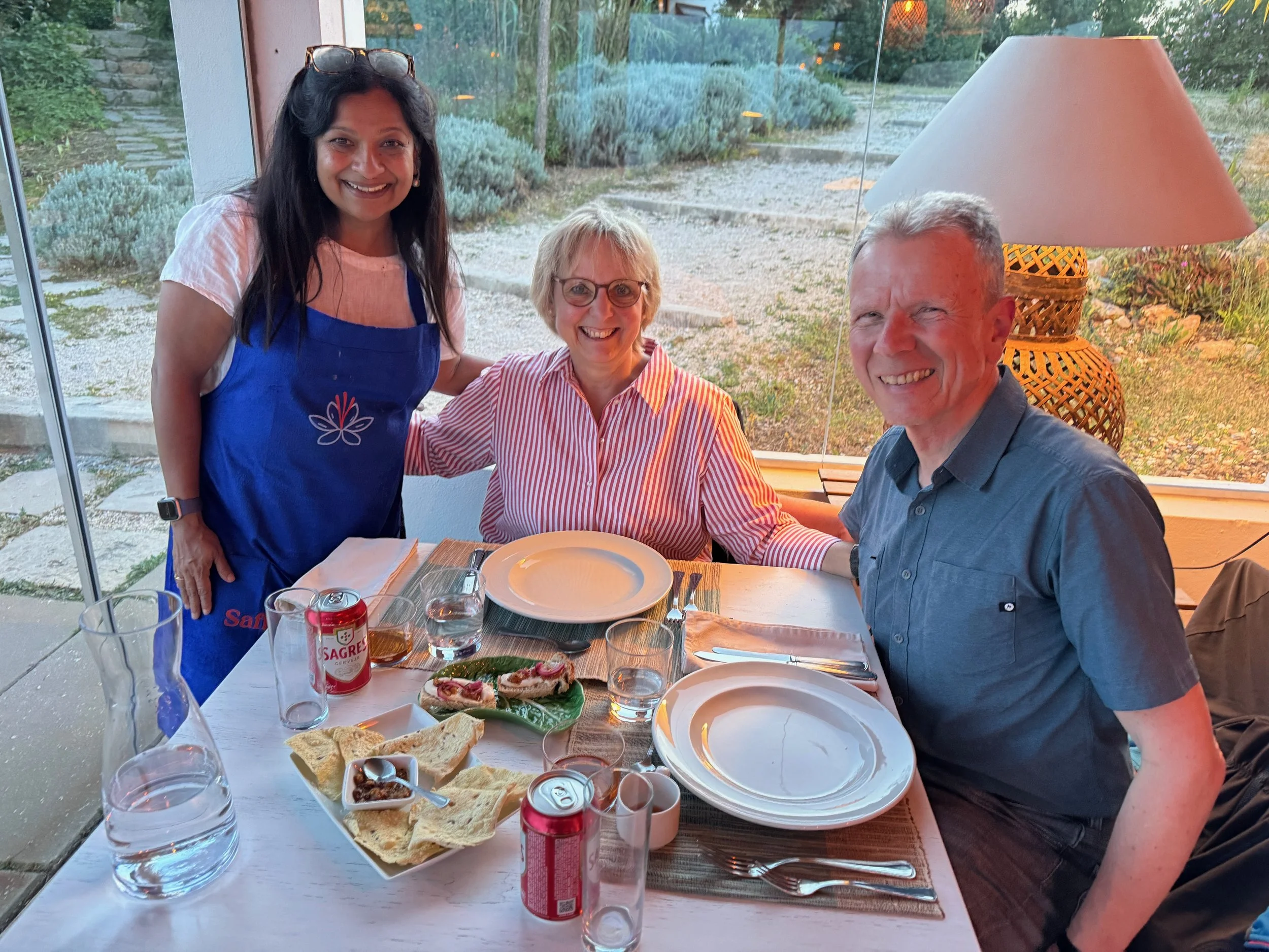 Three people smiling around a dining table with plates of food, beverages, and a basket of bread in a bright room with large windows overlooking a garden.