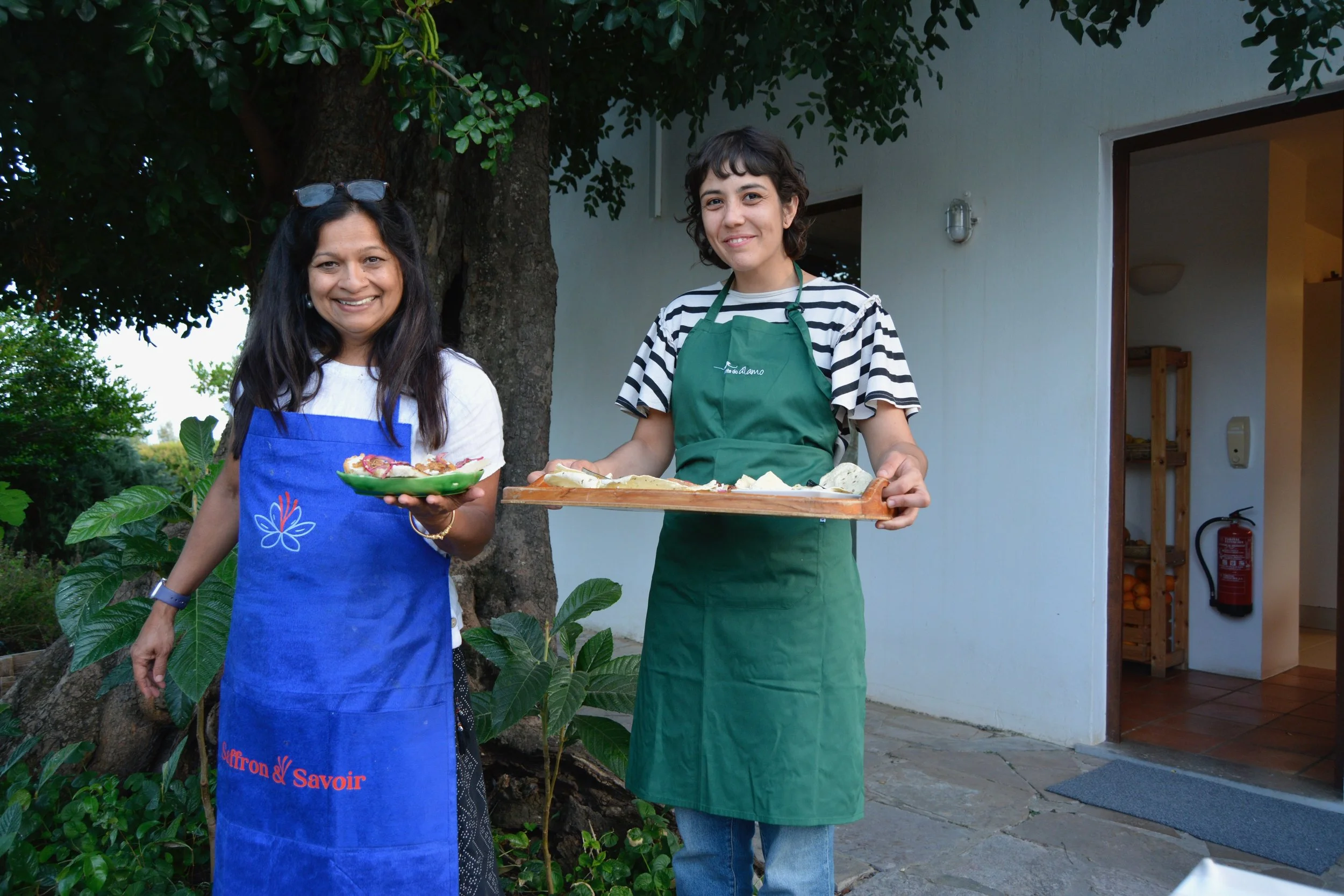 Two women standing outdoors in front of a tree and a white house, smiling at the camera. One woman is wearing a blue apron with red text and glasses on her head, holding a plate of food. The other woman is wearing a green apron over a black and white striped shirt and is holding a tray of food.