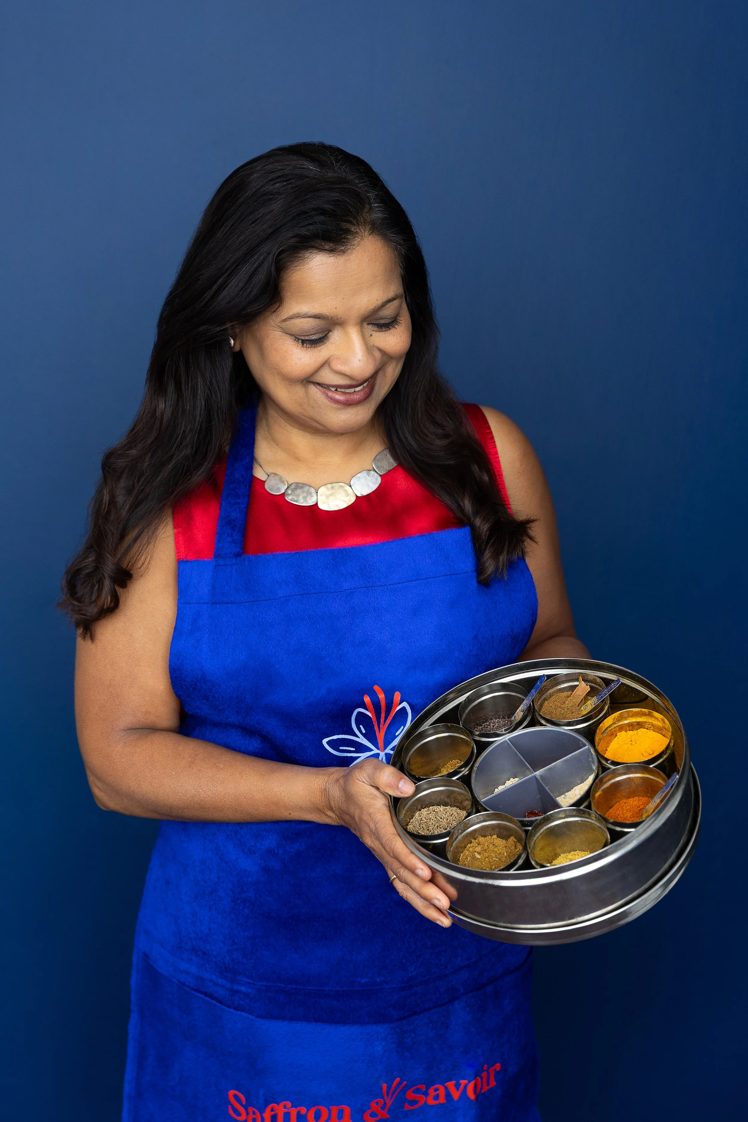 Indian Chef Sarita Bhagwat wearing a blue apron, smiling and holding a round container filled with various Indian spices.